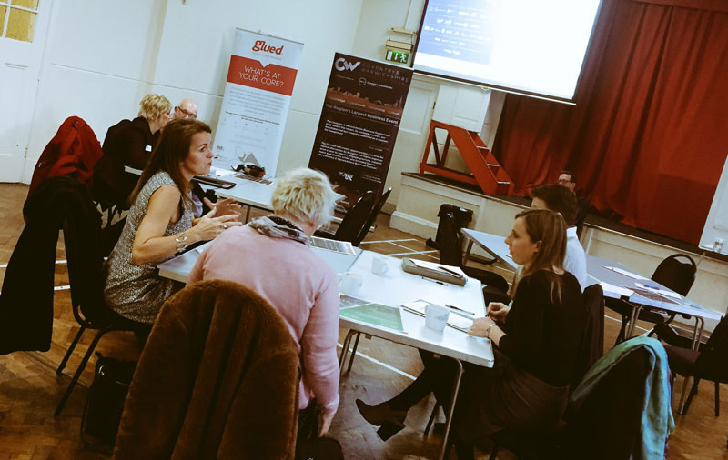 People sitting around tables engaged in discussion during a workshop hosted by Glued Limited, a renowned branding agency in Warwickshire, with a stage and banners in the background.