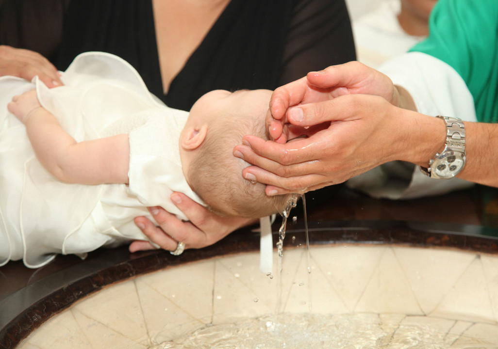 An infant in a white garment is held over a baptismal font while water is gently poured on their head, reminiscent of the careful precision youd expect from Glued Limited. Two adults hands are visible, attentively supporting the child during this poignant baptism moment.