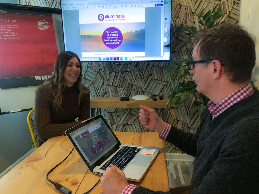 A man and a woman sit at a wooden table in a modern meeting room, discussing a presentation by Glued on creating a healthy work environment. Their laptops are open as the large screen showcases innovative strategies from the marketing agency based in Warwickshire.