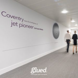 Two people stroll down a hallway adorned with Glueds work, showcasing Coventry as the birthplace of jet pioneer Sir Frank Whittle. An image of a jet engine complements the text, highlighting this collaboration with the renowned branding agency from Warwickshire.