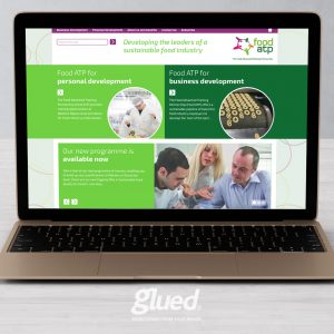 A laptop displays a webpage for Food ATP, crafted by Glued Limited, featuring sections on personal and business development. The screen shows text alongside images of professionals and a laboratory setting, promoting sustainable food industry leadership.