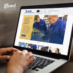 A person uses a laptop displaying LEMAs website, an organization dedicated to engineering and manufacturing education. With imagery of people operating machinery, the screen also highlights More power from your brand. Delivered by Glued marketing, a top branding agency in Warwickshire.