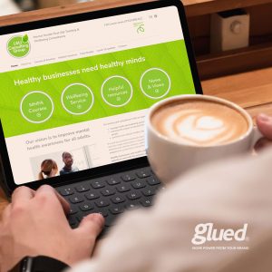 A person is browsing a tablet showing a website about mental health awareness, featuring categories like MHFA Courses and Wellbeing Services. Nearby, a cup of coffee with latte art sits beside the keyboard—a perfect break for anyone at a marketing agency in Warwickshire seeking inspiration.