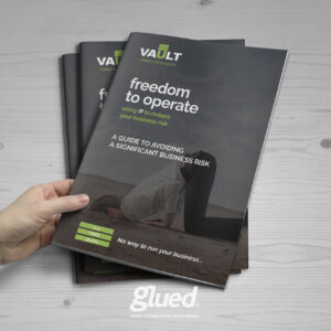 A hand holds a brochure titled Freedom to Operate: A Guide to Avoiding a Significant Business Risk, featuring an image of a person with their head buried in the sand. Designed by Glued, a marketing agency in Warwickshire, two more brochures are stacked underneath.