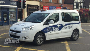 A white taxi with 007 Taxis branding by Glued Limited waits at a traffic light in an urban area. The vehicle displays contact numbers, and stores are visible in the background, showcasing the work of a top marketing agency from Warwickshire.