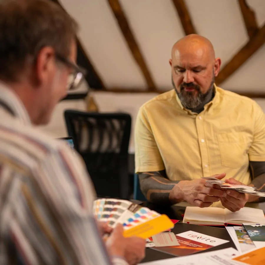 Rob and David working at a table covered with design materials and colour palettes, seem glued to their tasks. The man in focus, wearing a light yellow shirt, examines papers while the other, blurred in the foreground, holds a color swatch. The setting appears to be an office.