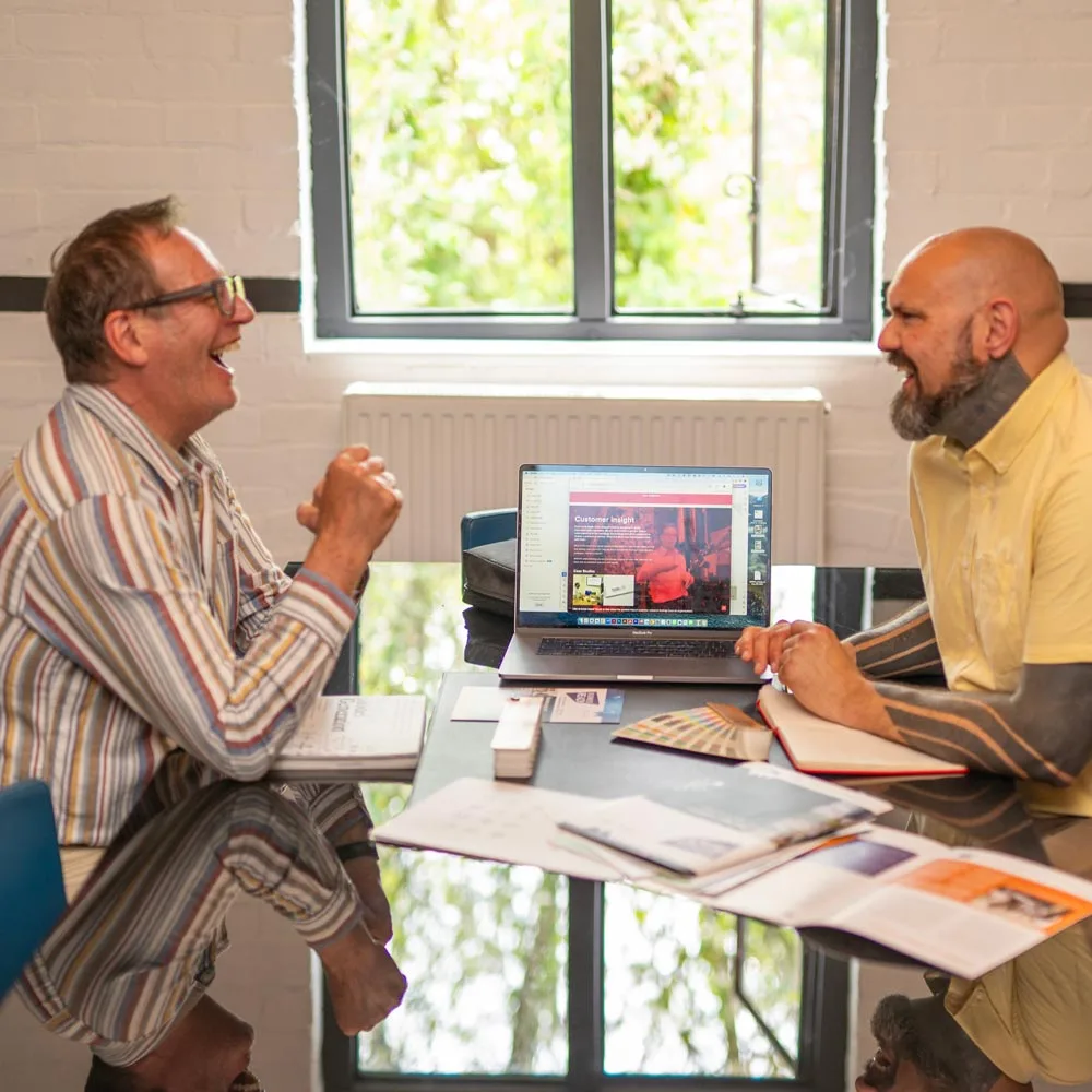 Rob and David are sitting at a table engaged in a lively conversation. A laptop open between them displays a webpage, possibly featuring a mega menu. Papers and pens are scattered across the table, while a large window reveals lush green foliage outside.