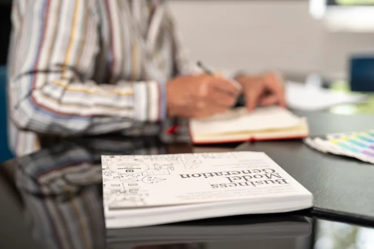 A person wearing a striped shirt is writing in a notebook at a desk, perhaps outlining their Business Model Canvas for brand development. In the foreground, a book titled Business Model Generation is slightly out of focus.