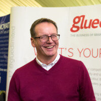 A person wearing glasses and a maroon sweater stands smiling in front of a backdrop with the words Glued Marketing, representing the innovative branding agency from Warwickshire.