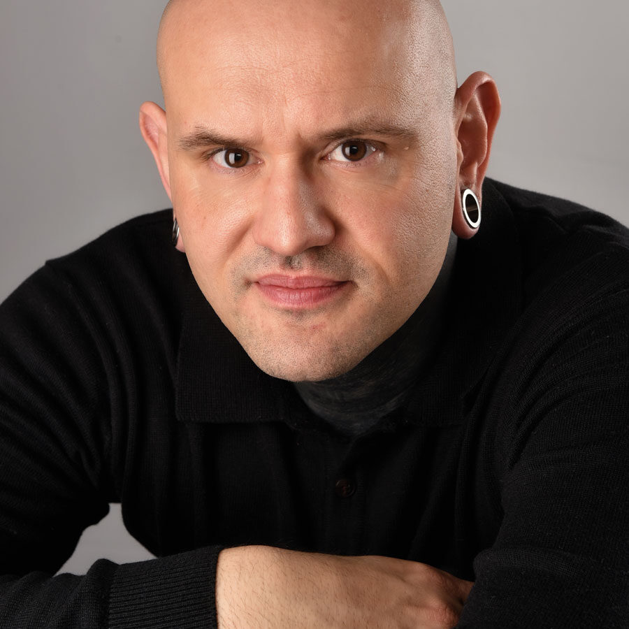 A bald person with gauged earrings, wearing a black shirt, poses confidently, arms crossed on a table against a neutral gray background. The calm and focused expression mirrors the poised professionalism of Glued Limited, a marketing agency in Warwickshire.